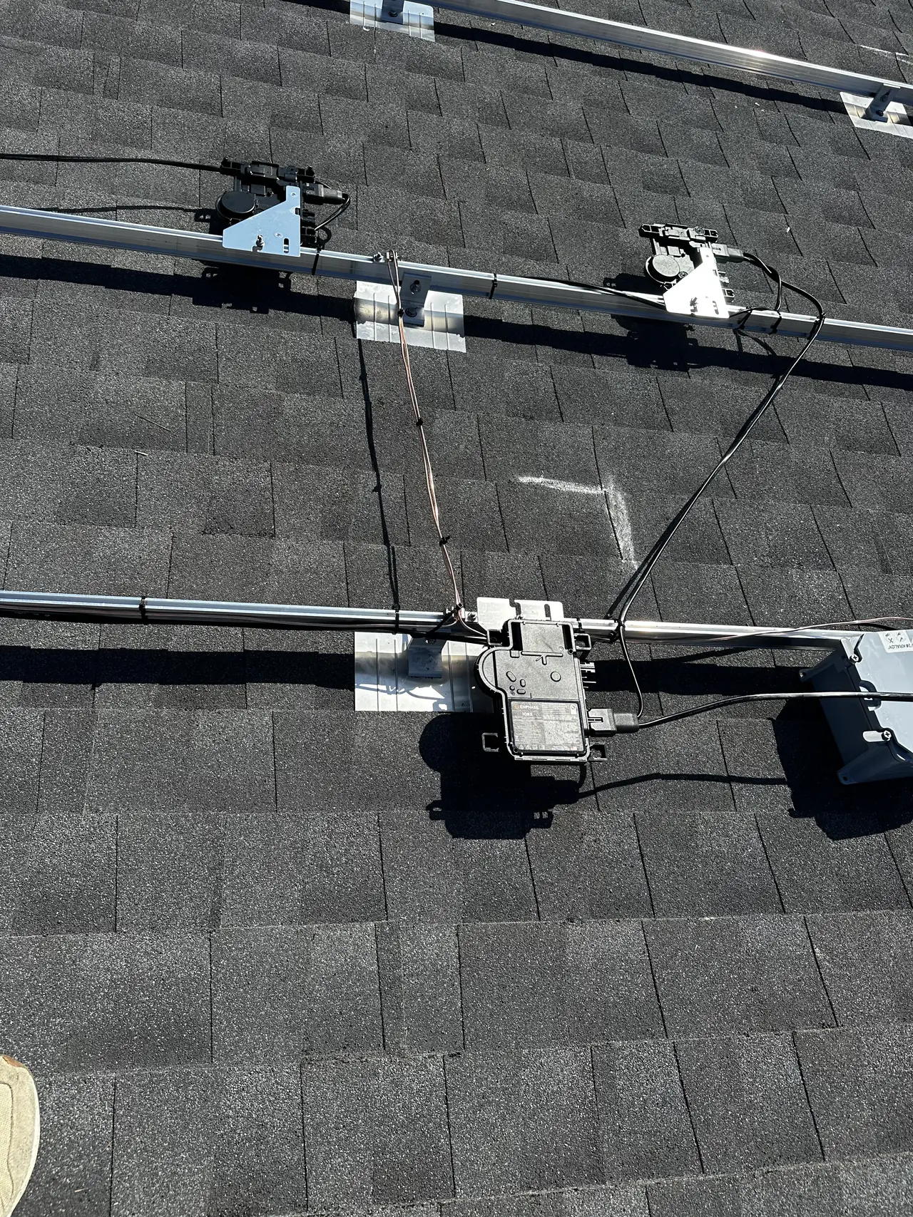 A microinverter clipped to a mounting rail on a shingle roof, with the trunk cable and a bare bonding conductor dressed along the rail and bright metal flashings under the feet, Studio City