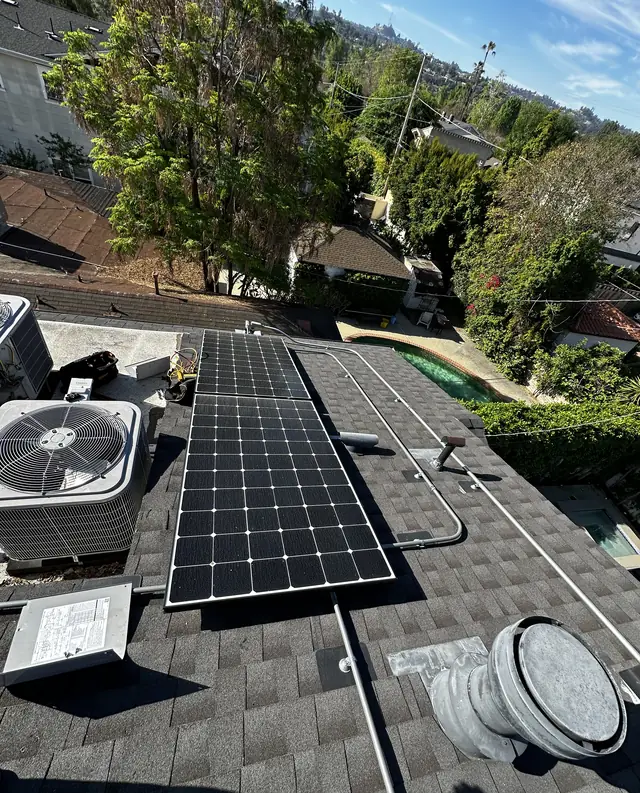 Solar modules on a shingle roof beside rooftop air-conditioning condensers, with trees and neighboring rooftops beyond, Studio City