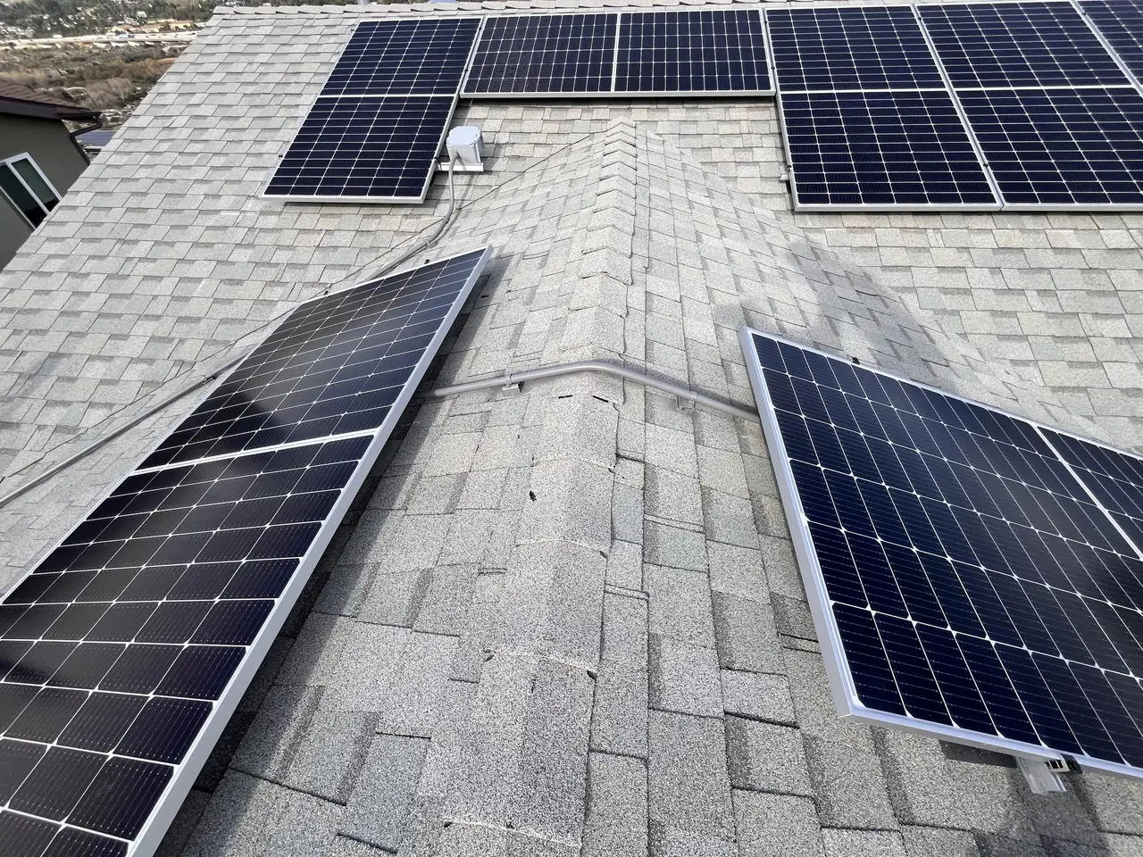 Two groups of solar modules on either side of a shingle roof ridge, joined by a conduit over the ridge, Sunland