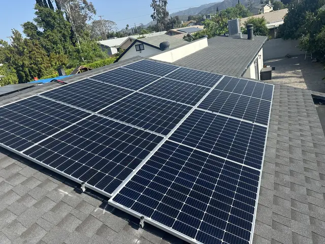 Rows of solar modules on a dark shingle roof with neighboring houses and foothills behind, Sunland