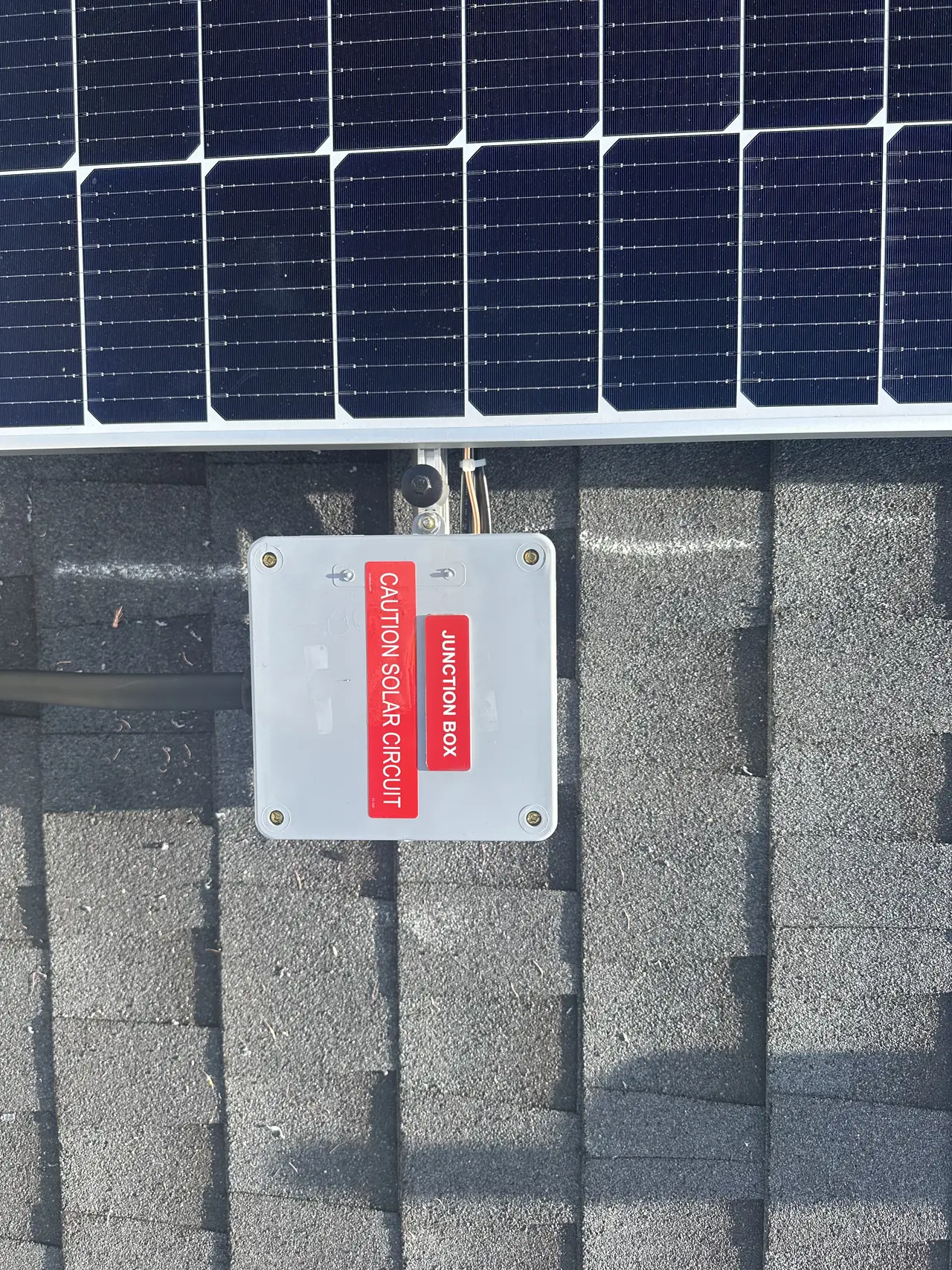 A closed junction box on a shingle roof labeled JUNCTION BOX and CAUTION SOLAR CIRCUIT, beside the array, Sunland