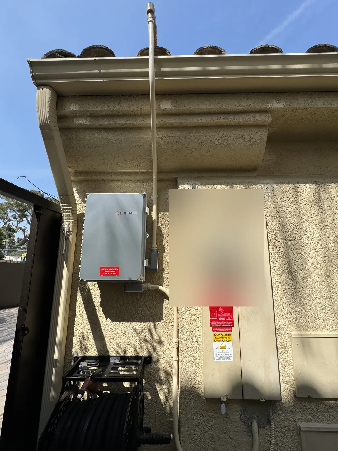 Enphase combiner and meter panel side by side under the eave of a stucco wall, with solar warning labels, the meter face obscured, Tarzana