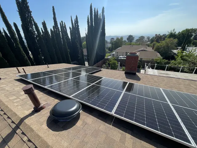 Solar modules on a brown shingle roof beside a brick chimney, with cypress trees and foothills beyond, Tujunga