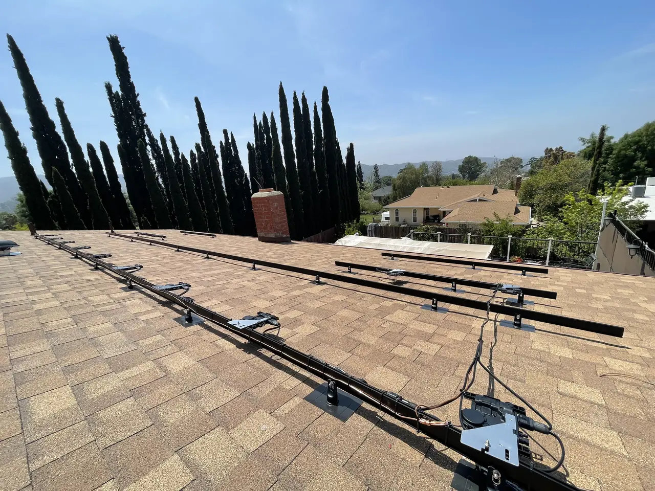 Black rails with Enphase microinverters on a shingle roof before the modules, cypress trees behind, Tujunga