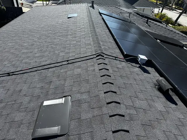 Black solar modules along a ridge on a dark shingle roof with a roof vent in front, Tujunga