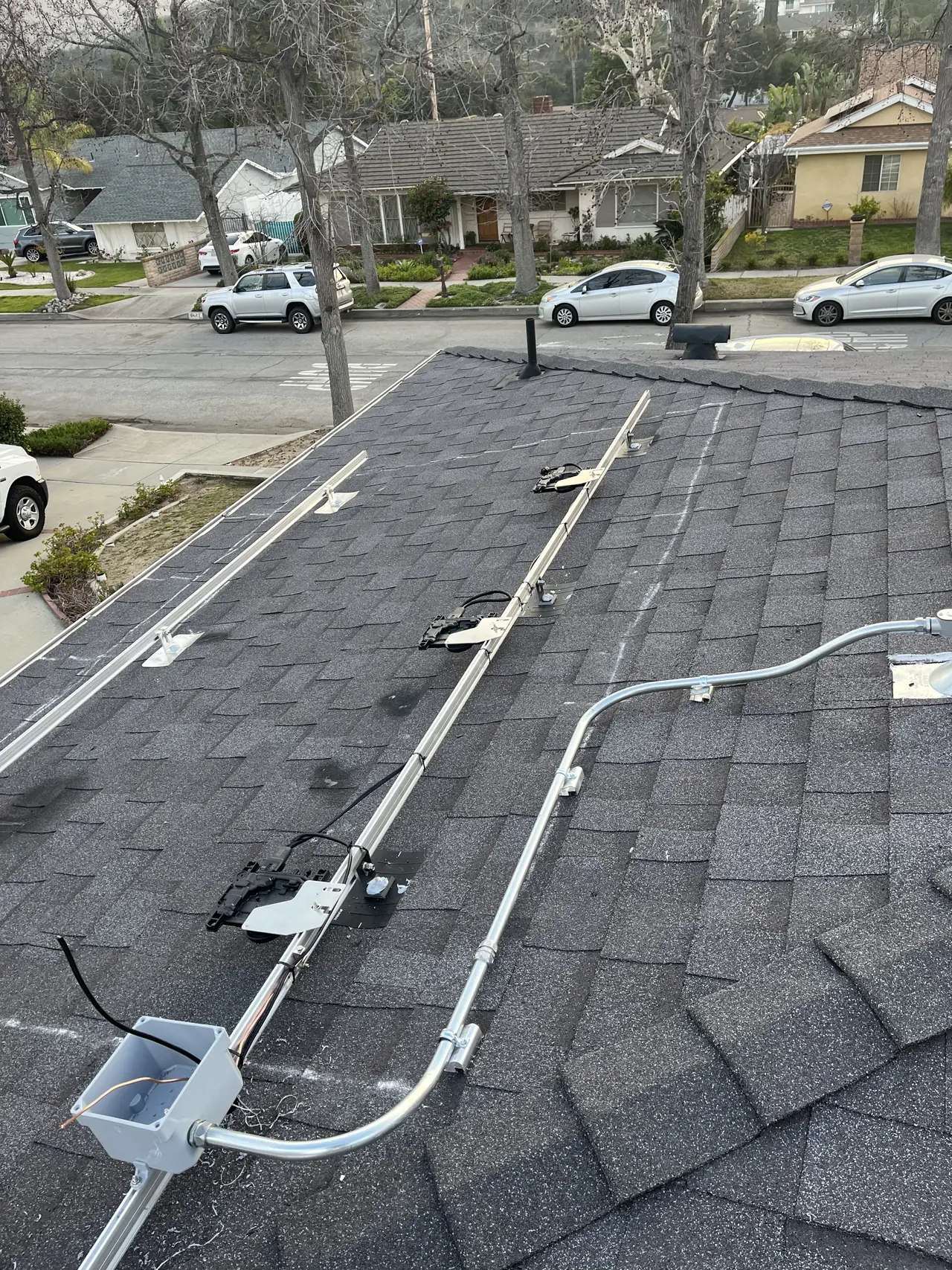 Rails with Enphase microinverters and a junction box on a dark shingle roof before the modules, Tujunga