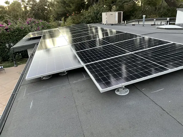 Solar modules on tilt legs over a low-slope roof, with trees and flowering shrubs beyond, Tujunga