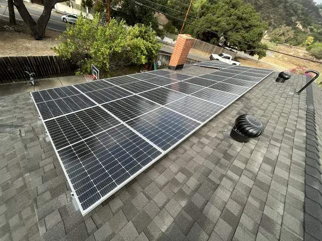 Solar modules on a dark shingle roof with a turbine vent, a chimney and trees beyond, Tujunga