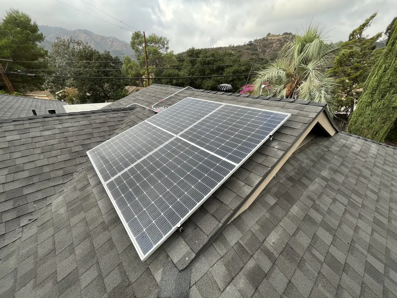 A small solar array by a gable on a dark shingle roof with wooded hills behind, Tujunga