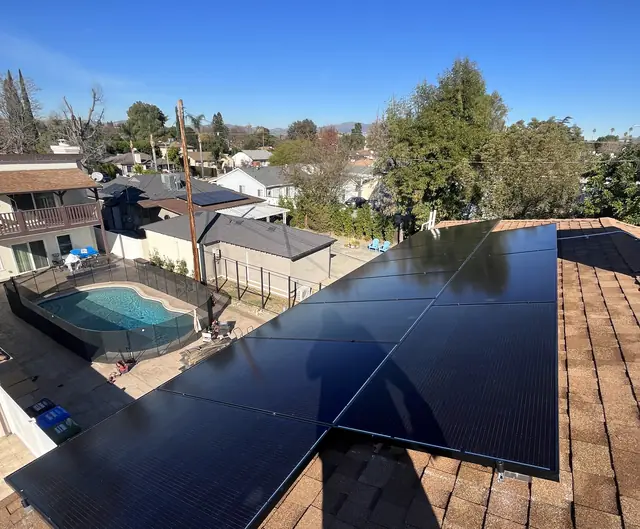 Black solar modules in two rows meeting at the ridge of a brown shingle roof, with a pool and neighboring houses beyond, Van Nuys, Los Angeles