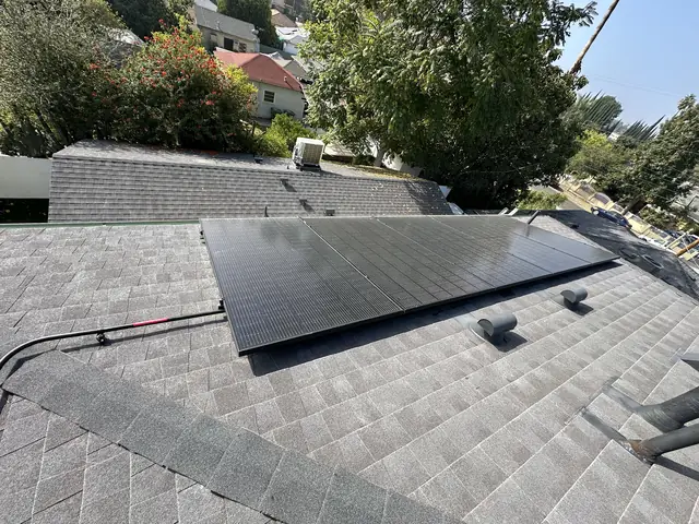 Black solar modules on a dark gray shingle hip roof with trees and rooftops beyond, Van Nuys, Los Angeles