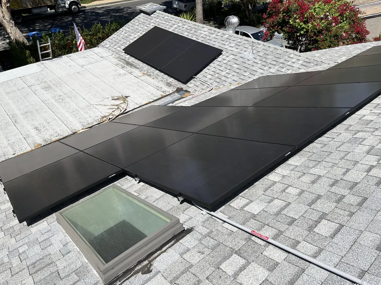 A group of black modules beside a skylight on a light-colored shingle roof with trees and rooftops behind, Van Nuys, Los Angeles