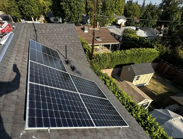 A short row of black solar modules on a dark charcoal shingle roof with a garden and hedge below, Van Nuys, Los Angeles