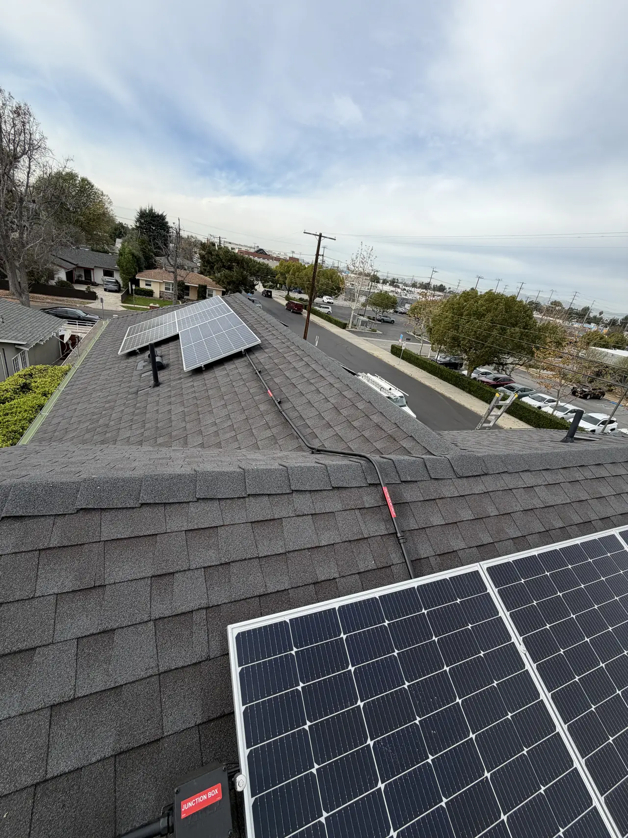 Black modules in a short row on a dark shingle roof with power lines and rooftops behind, Van Nuys, Los Angeles