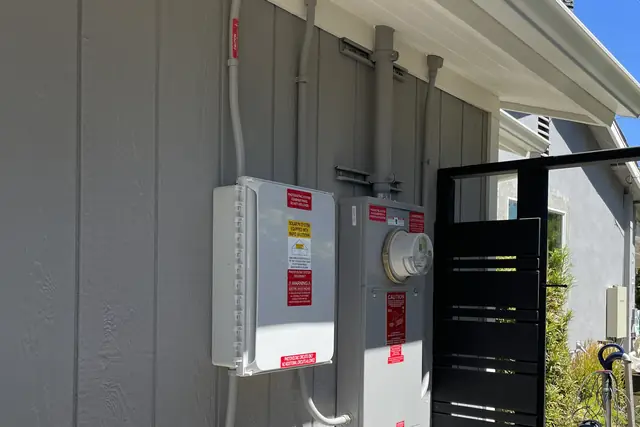 Solar disconnect and meter panel mounted on the gray board-and-batten side wall of a house under a clear sky, Woodland Hills