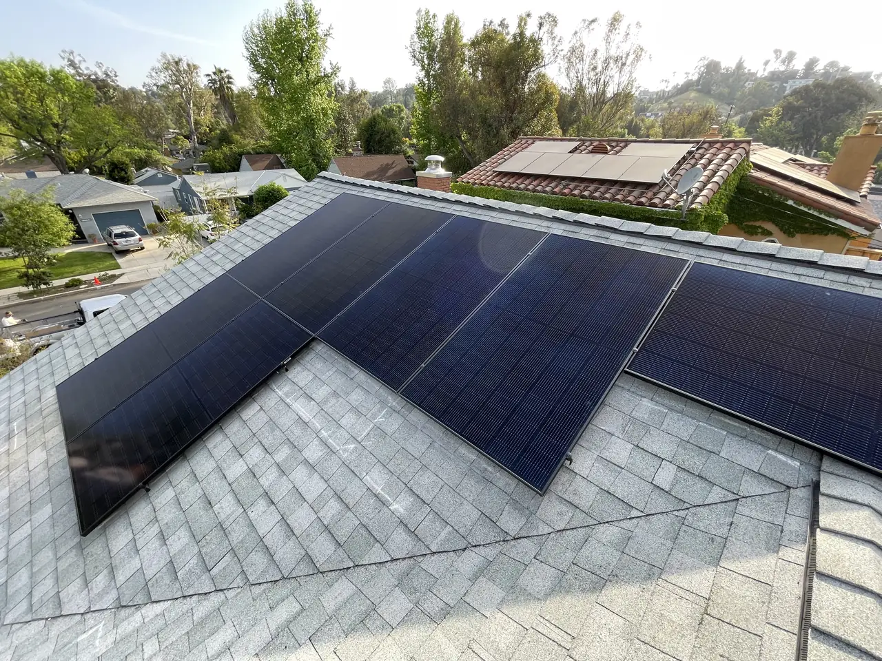 Two sections of black solar modules meeting at the hip of a new light gray shingle roof with the neighborhood beyond, Woodland Hills