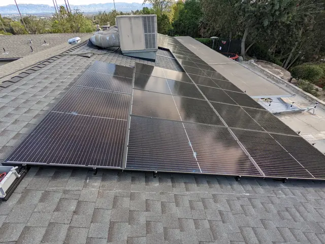 A large black solar array covering several planes of a gray shingle roof with trees and the valley beyond, Woodland Hills