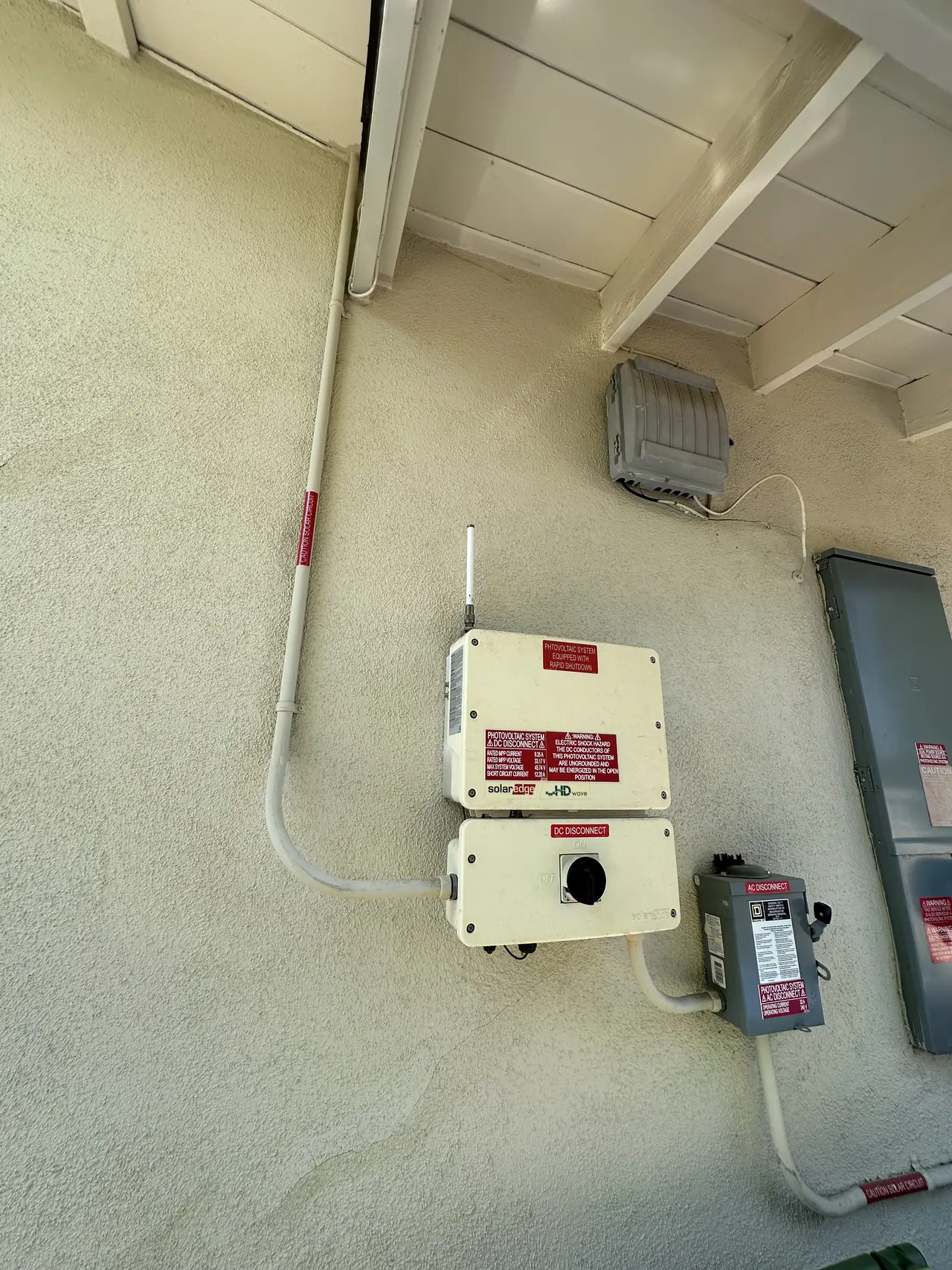 An equipment wall with the inverter, disconnect and conduit runs on stucco beneath a wooden eave, Woodland Hills