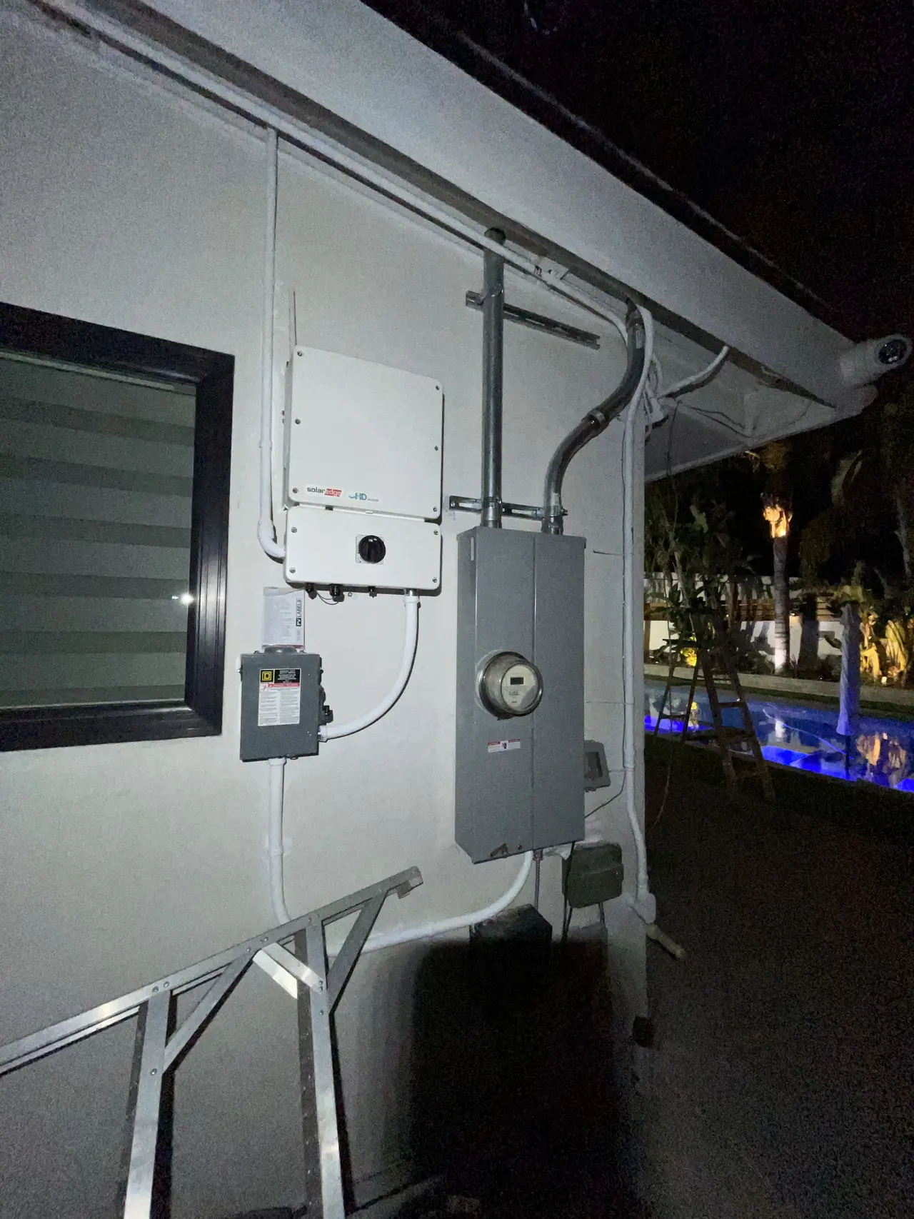 An inverter, a disconnect and a meter panel mounted along an exterior wall, all closed, photographed in the evening, Woodland Hills